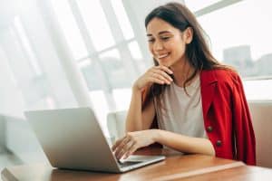 cheerful attractive young woman working in cafe. look on laptop and smile. wear red jacket. fashionable model typing on keyboard. alone in room.