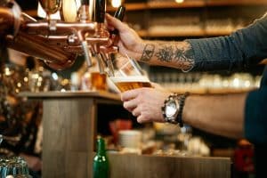 tattooed caucasian barman pouring beer while standing in pub. selective focus on hand.